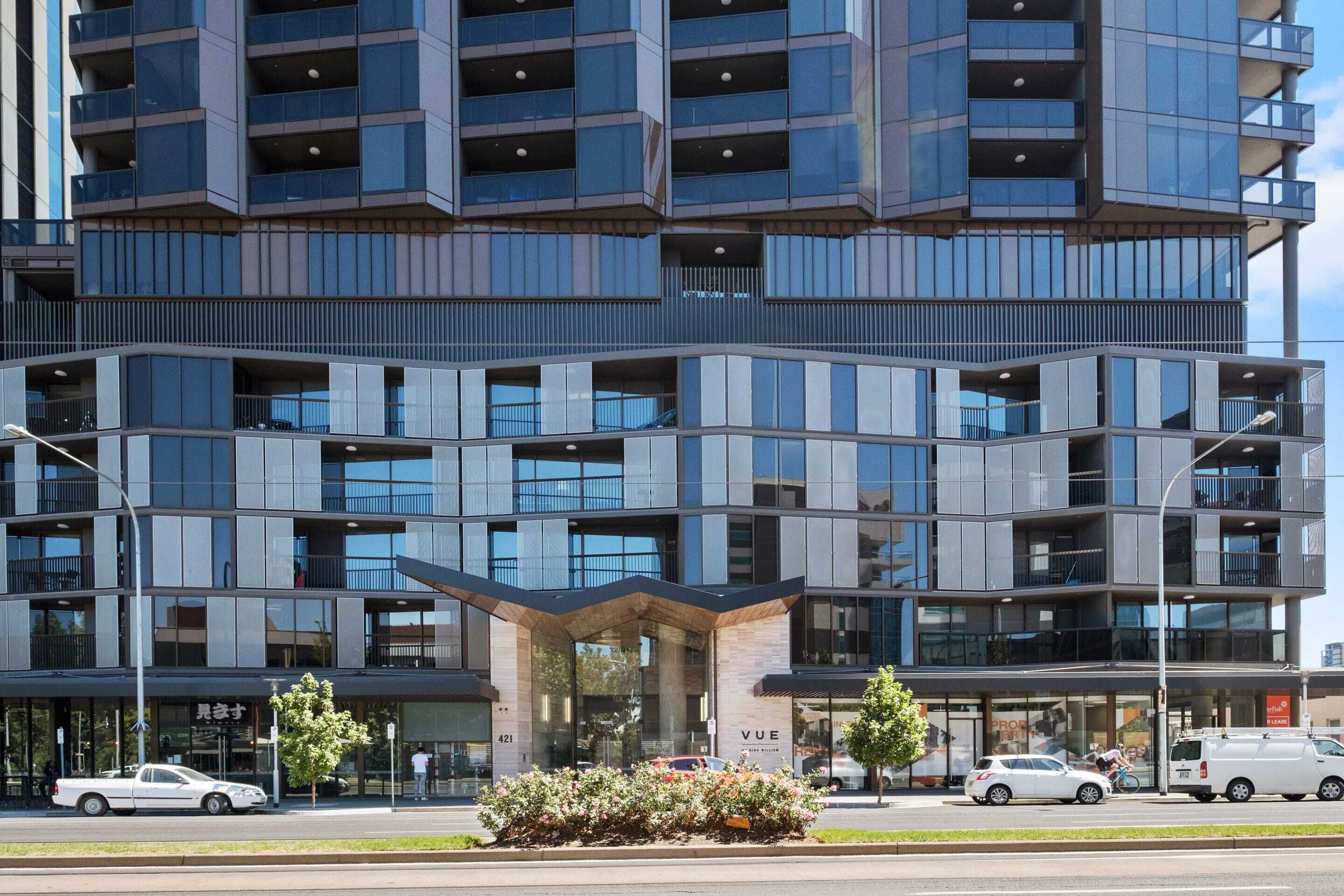 A modern multi-story building with a prominent entrance, large windows, and a landscaped area with flowers.