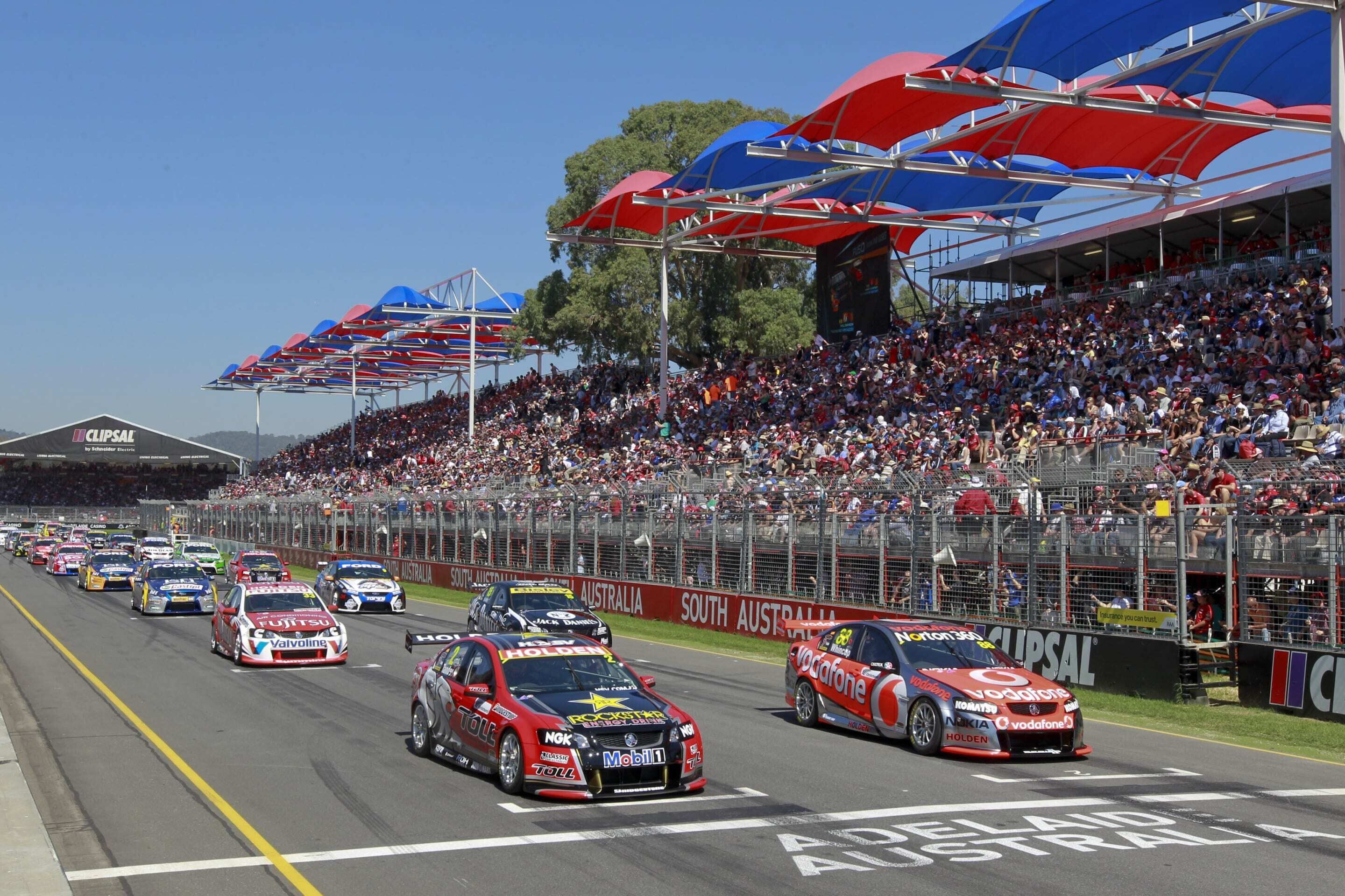 A racing event with multiple cars on a track, spectators in the stands, and a clear sky.