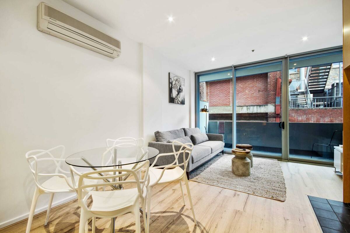 A modern living room with a glass table, white chairs, a grey sofa, and a large sliding glass door leading to an outdoor area with a brick wall.