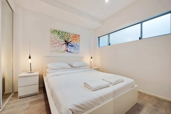 Een moderne slaapkamer met een groot bed, wit beddengoed, twee nachtkastjes met lampen, een kleurrijk schilderij van een boom aan de muur, en een raam met zonwering.