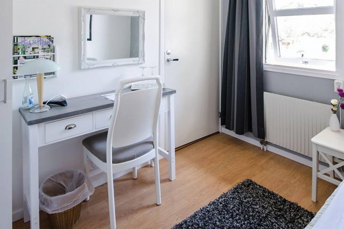 Une chambre à coucher comprenant un lit, un bureau, une chaise, un radiateur, une fenêtre avec des rideaux et une petite poubelle.