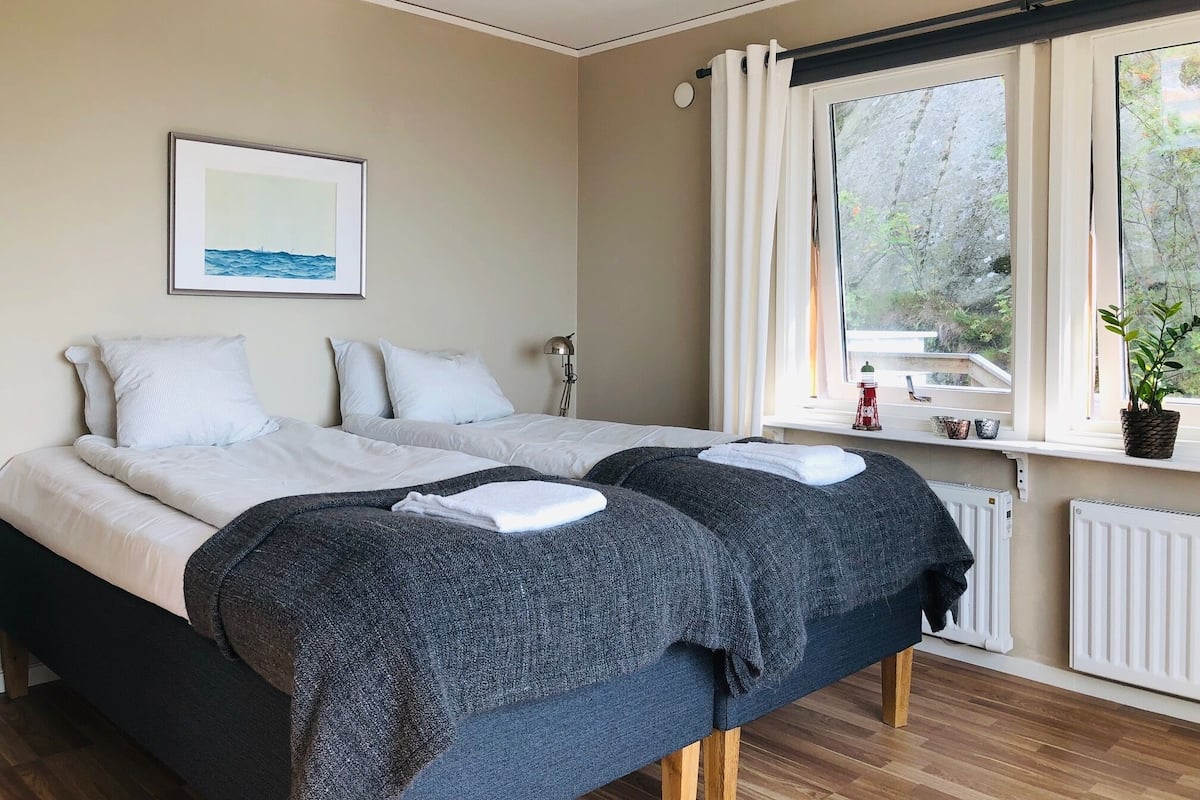 Une chambre avec deux lits, un tableau encadré au mur, une fenêtre avec des rideaux, un radiateur et une plante sur le rebord de la fenêtre.