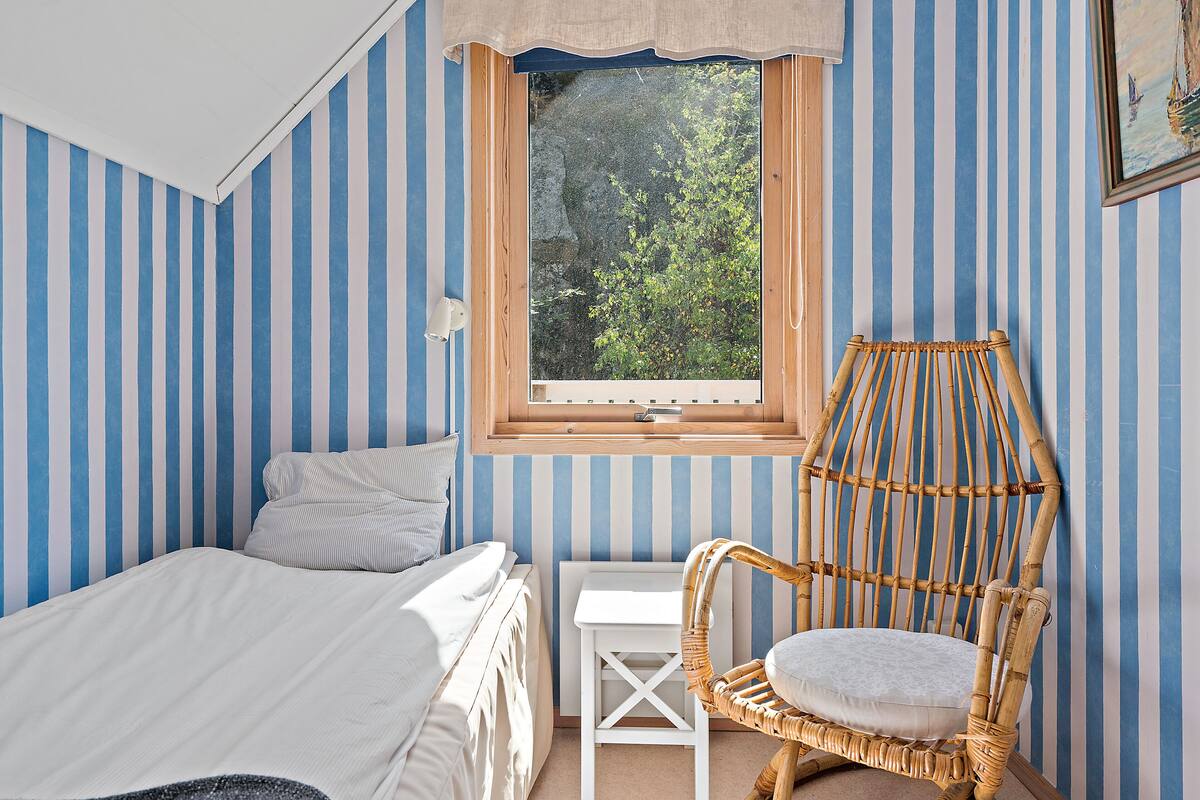 Une chambre à coucher avec un lit, une petite table, une chaise en osier et une fenêtre donnant sur des arbres.