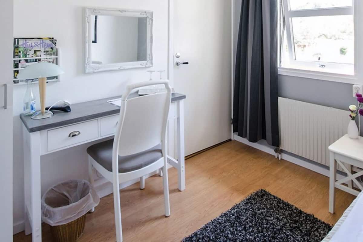 Une chambre à coucher comprenant un lit, un bureau avec une chaise, une fenêtre avec des rideaux, un radiateur et une petite poubelle.
