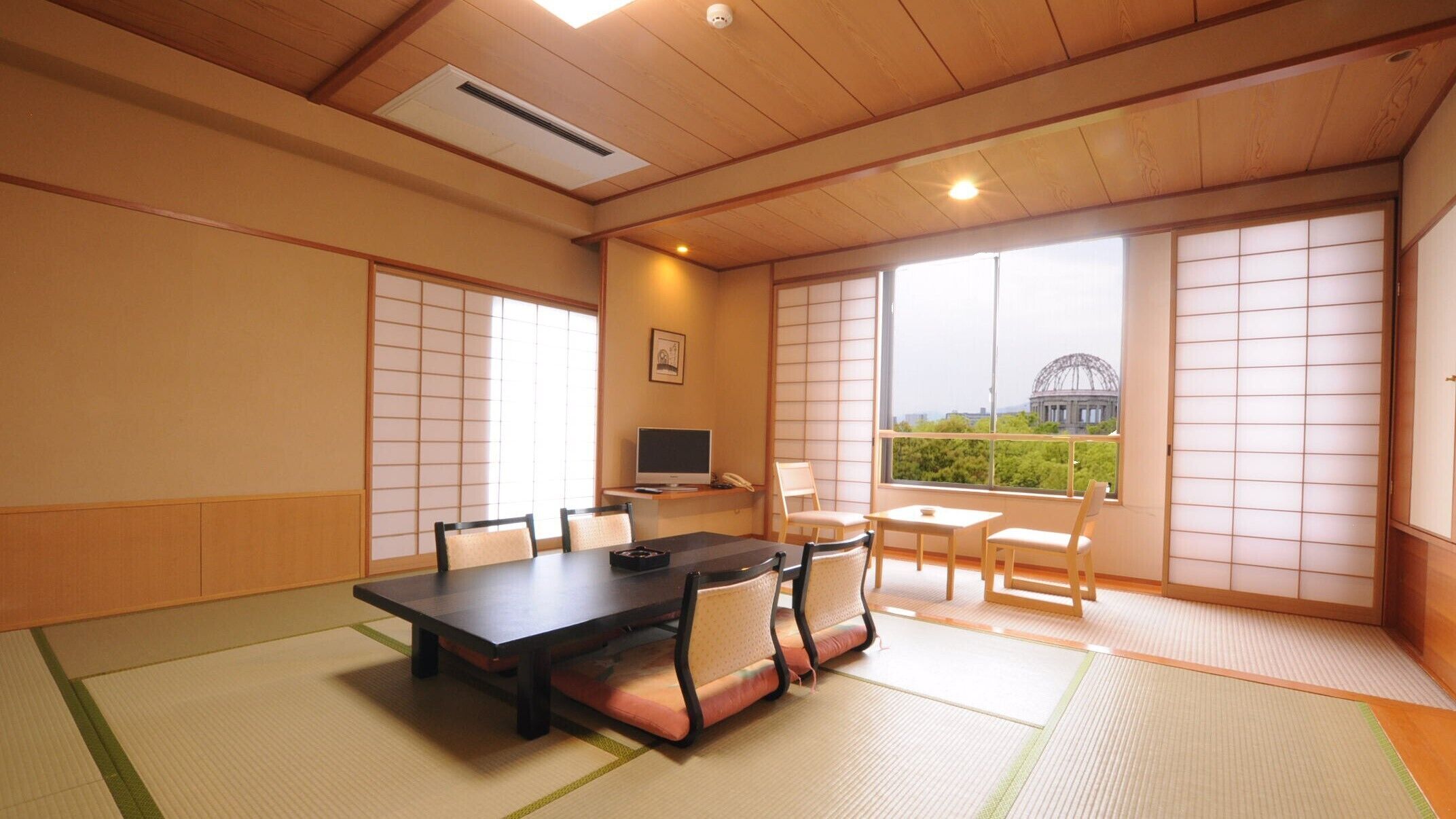 A traditional Japanese-style room with tatami flooring, a tatami mat border, a wooden table, chairs, a television, and large windows with a view of a building.