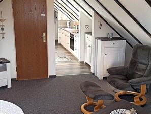 Um interior moderno com porta de madeira, uma cadeira estofada cinza, um banco de madeira e uma área de cozinha com armários brancos e bancada preta.