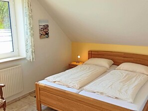 A bedroom with a wooden bed, two pillows, a nightstand with a lamp, a window with curtains, and a radiator.