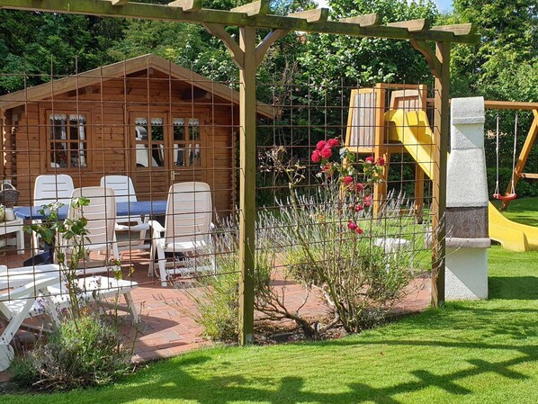 A wooden cabin with a patio area, a yellow swing set, and a garden with various plants.