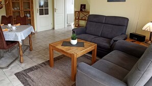 A living room with a sofa, a dining table with chairs, a wooden coffee table, and a cabinet with glass doors. - Apartment Lee - Apartments on the old dike in the guest house (Wittmund)