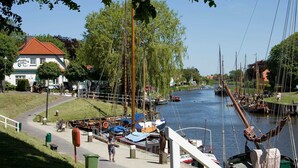 A riverside view with a restaurant, boats docked along the water, and a pedestrian walkway. - Apartment Lee - Apartments on the old dike in the guest house (Wittmund)