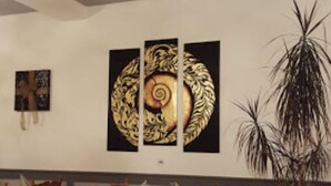 Een eetgedeelte met houten stoelen en tafels, een grote potplant en een wand met drie ingelijste kunstwerken.