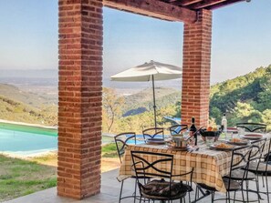 Un espace extérieur abrité, aménagé pour les repas, avec des piliers en briques, une table dressée avec des assiettes, des verres et une bouteille, offrant une vue panoramique sur un paysage pittoresque avec une piscine et des collines.