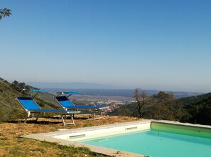Une piscine avec deux chaises longues bleues, entourée d’un paysage vallonné et sous un ciel dégagé.