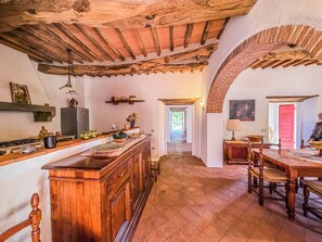 A rustic kitchen with wooden beams, a brick floor, and a dining area with a table and chairs.