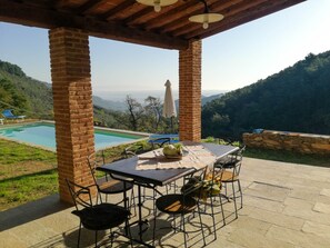 A covered outdoor seating area with a table and chairs, overlooking a pool and a mountainous landscape.