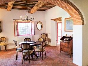 A dining area with a round table and six chairs, a wooden dresser, a chandelier, a window with a view, and a mirror on the wall.