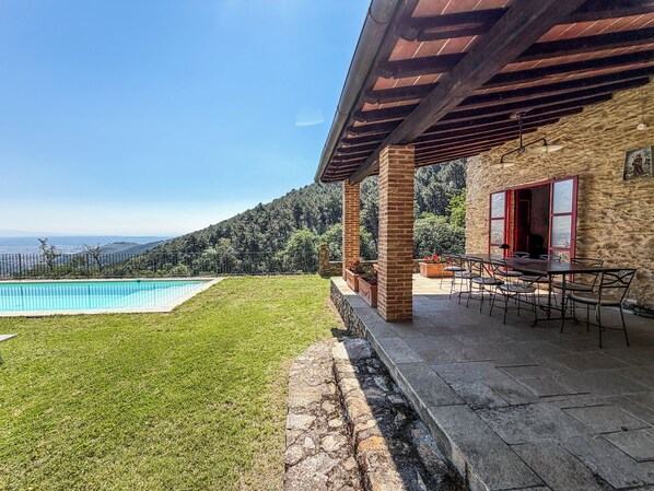 A poolside area with a view of the sea and hills, a covered patio with a table and chairs, and a stone wall.