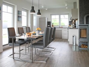 A modern kitchen with a dining area, featuring a long table with chairs, pendant lights, and a view of the outdoors.