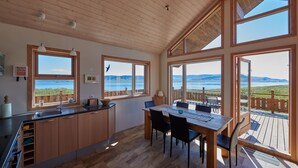 A spacious kitchen with wooden cabinets, a dining area with a table and chairs, large windows with a view of the sea, and a wooden deck. - Brekka - Stunning Country & Ocean View Location, Yet Close To Reykjavik (Akranes)