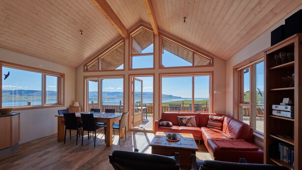 A spacious living room with a large wooden ceiling, a red sofa, a dining area with a table and chairs, and large windows offering a view of the landscape. - Brekka - Stunning Country & Ocean View Location, Yet Close To Reykjavik (Akranes)