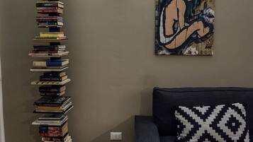 Una sala de estar con una alta pila de libros contra la pared, un sofá con un cojín estampado y una obra de arte colgada en la pared.