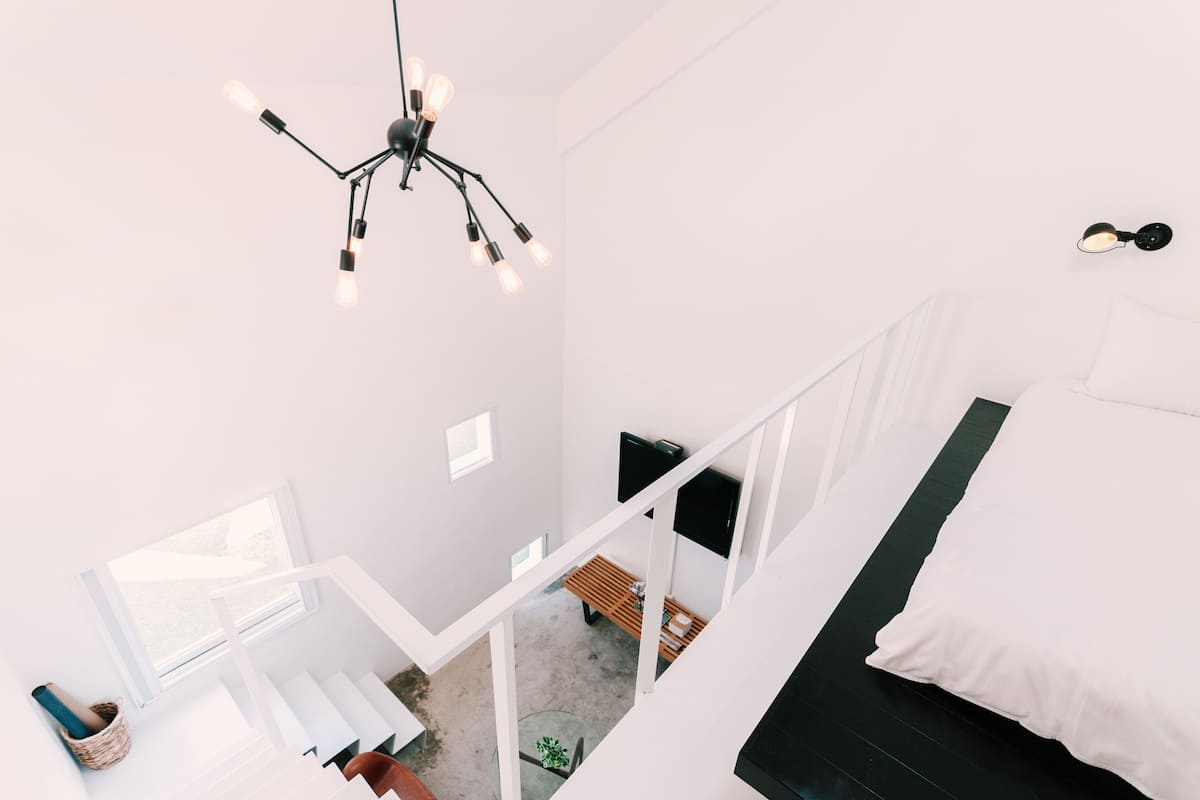 A high-ceiling room with a modern chandelier, a bed with white bedding, a staircase, and a view of the outdoors.