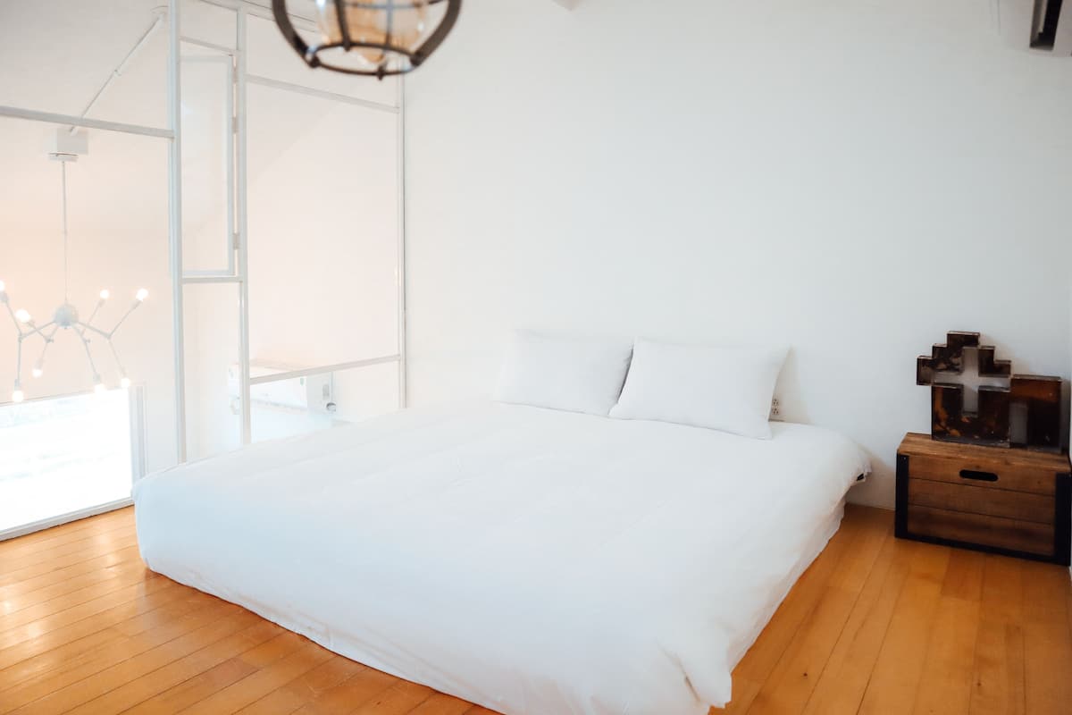 A bedroom with a large bed, wooden flooring, a nightstand with a lamp, and a view of the outdoors through a large window.