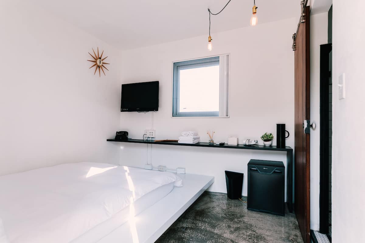 A modern bedroom with a bed, a TV mounted on the wall, a window, a small shelf with toiletries, and a door.