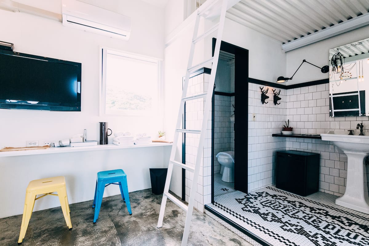 A modern bathroom with a white tiled wall, a black toilet, a white sink, a black trash bin, a white ladder, and a black and white patterned rug.