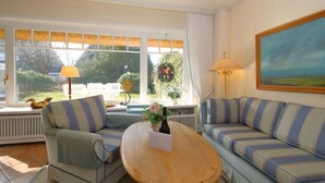 A living room with a sofa, armchair, and a round wooden coffee table. There is a window with a view of greenery and a painting on the wall. - House Tondernhüs, Germany (Sylt-Ost)