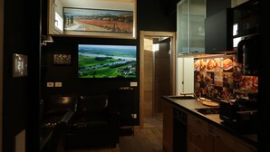 A modern kitchen with a built-in oven, a coffee machine, and a refrigerator. There is a television mounted above a leather couch and a wooden cabinet. - Michele Guest House (Pisa)
