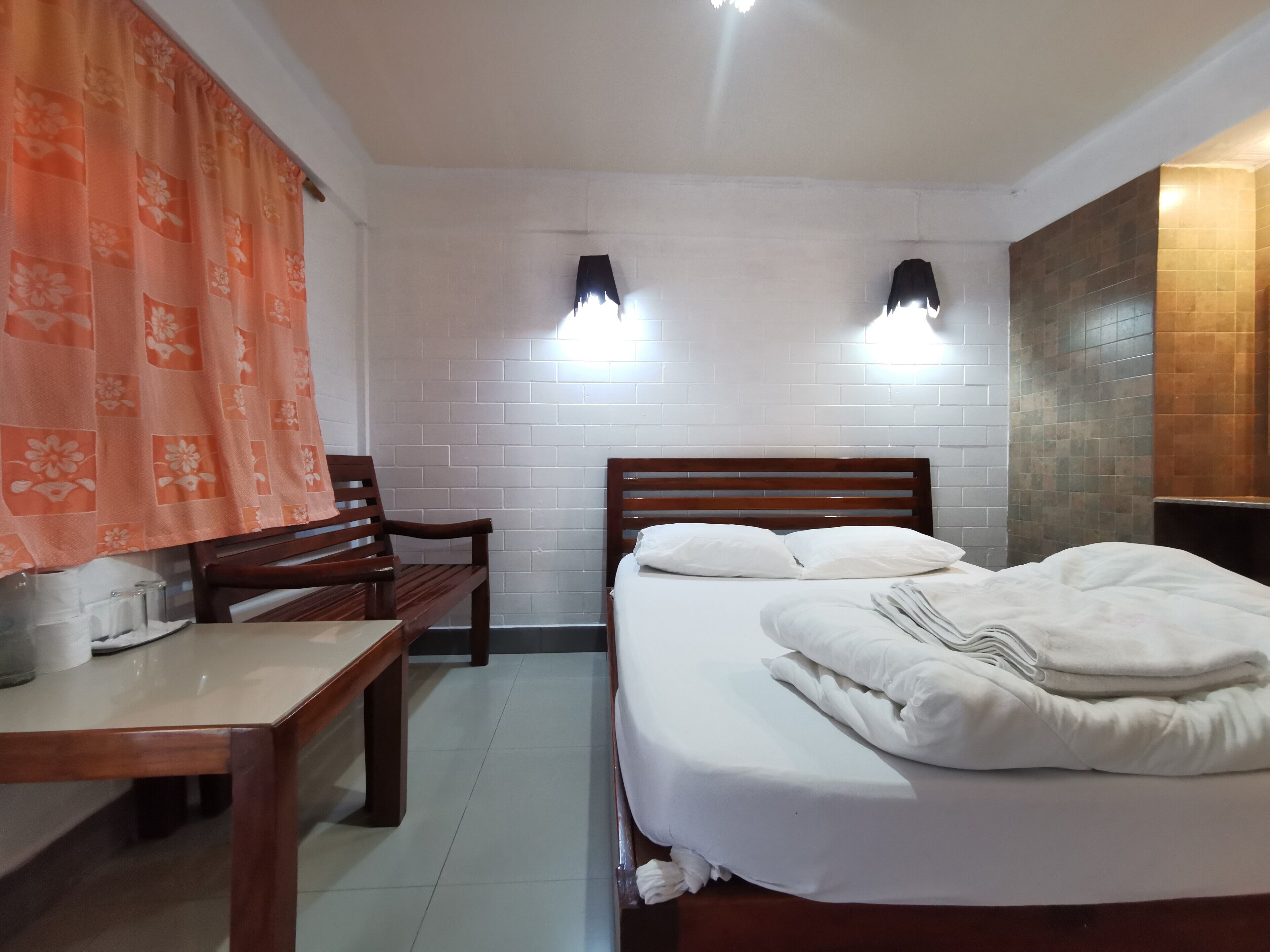 A hotel room with a wooden bed, white pillows, a wooden bench, a desk with a lamp, and a white brick wall.