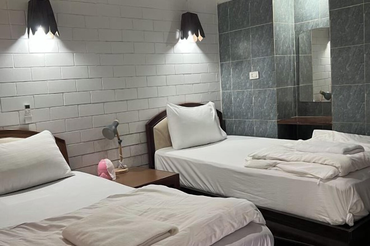 A hotel room with two beds, a nightstand with a lamp, a mirror, and a brick-patterned wall.