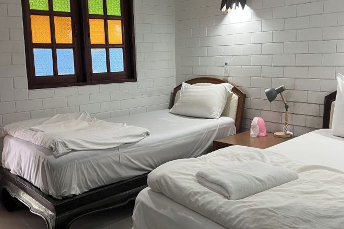 Quarto com duas camas, uma janela com vidros coloridos, uma mesinha de cabeceira com abajur e uma parede de tijolos.