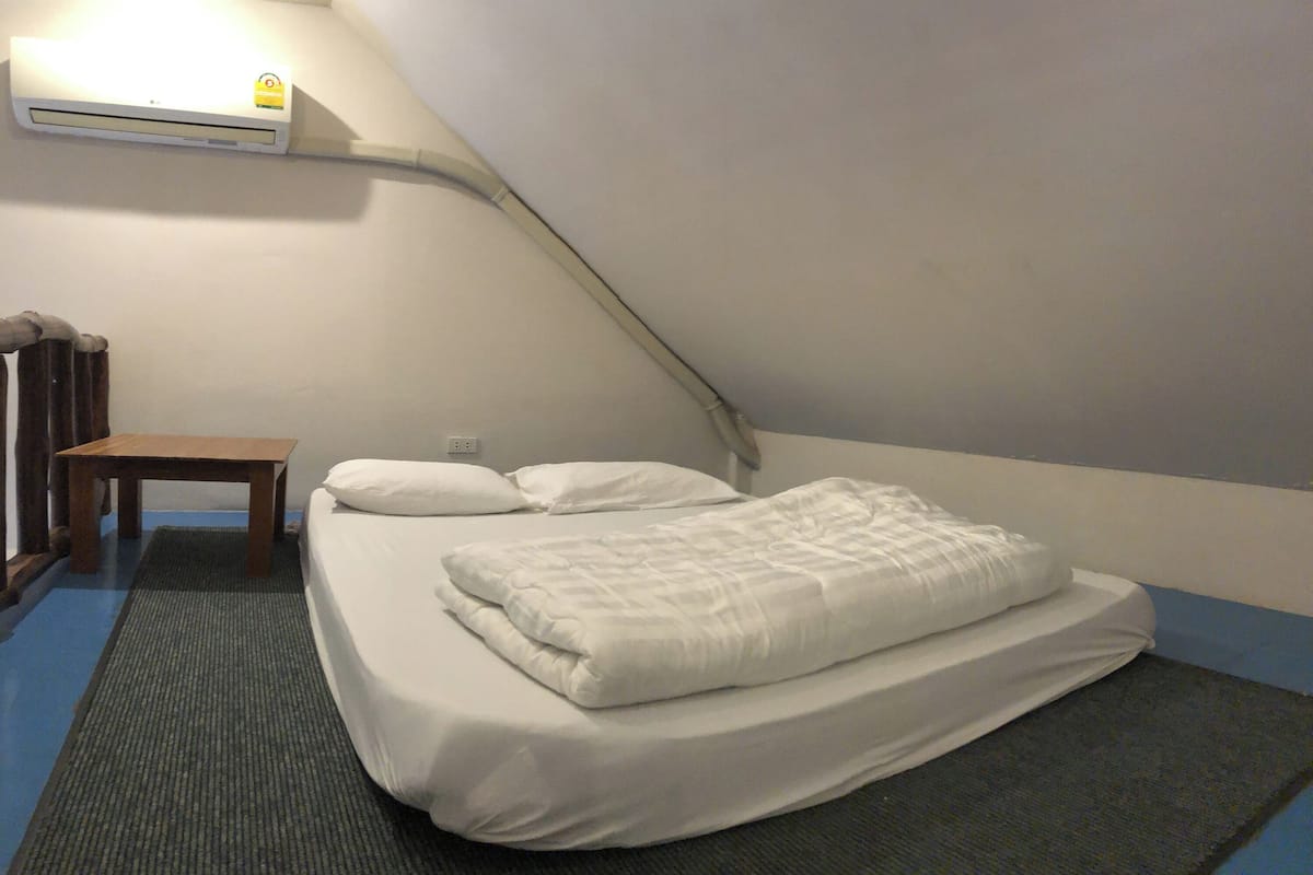 Een eenpersoonsbed met witte beddengoed in een kamer met een schuine kap en een airconditioner aan de muur.
