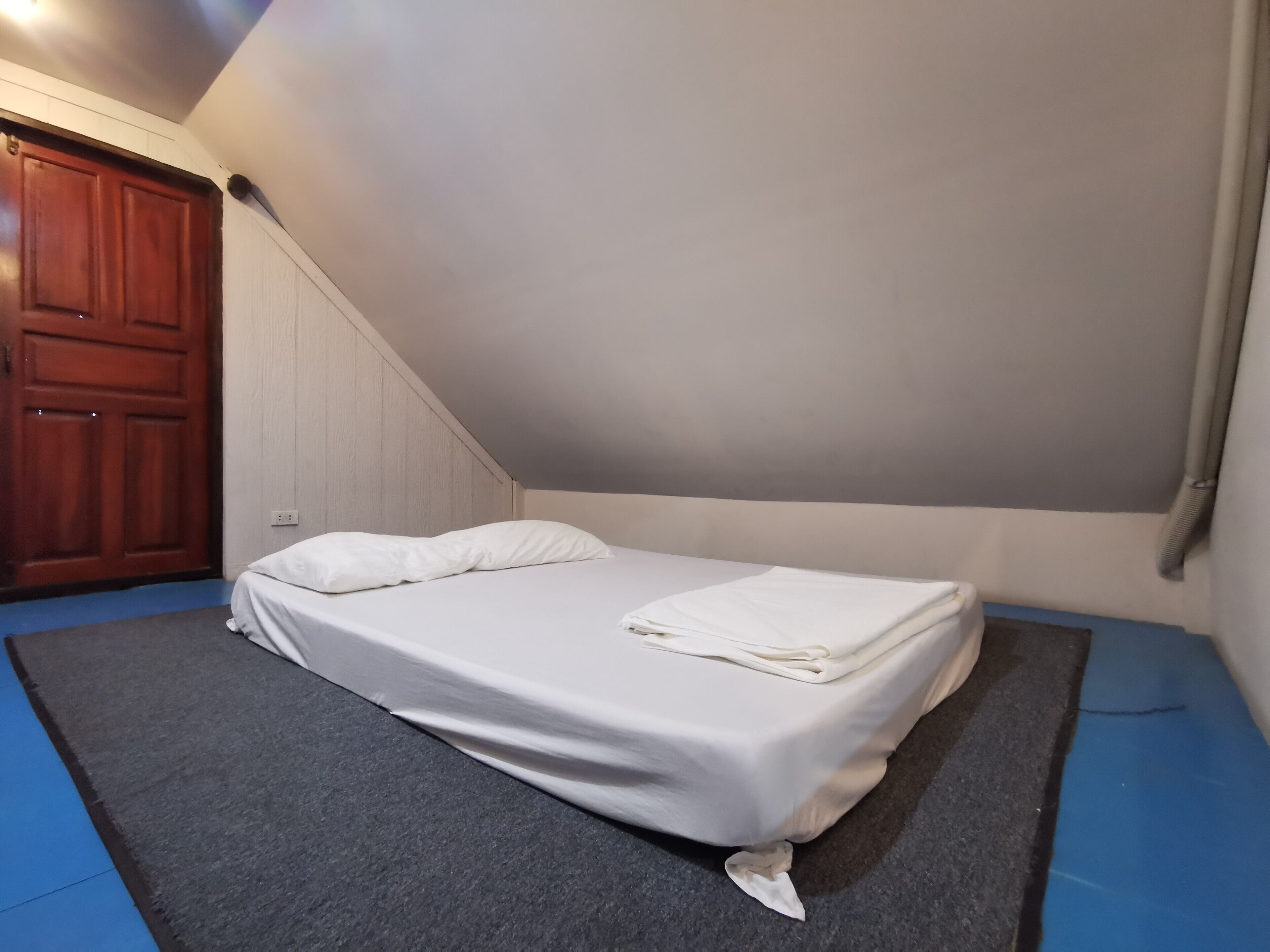 Quarto pequeno com cama de solteiro, porta de madeira, parede branca e teto inclinado.