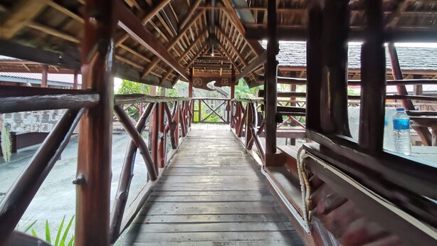 A wooden bridge with a metal railing, leading to a covered area with a tiled floor and a view of greenery outside. - Baan Lanna Resort (Chonburi)