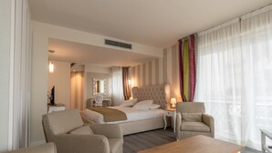 A hotel room with a bed, two armchairs, a wooden coffee table, a side table, a mirror, and a window with sheer curtains. - La Dolce Vita Hotel (Menton)