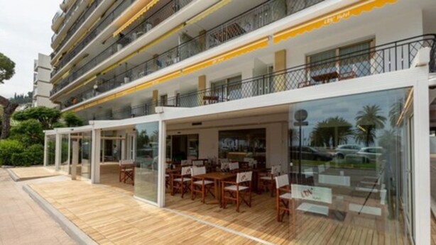 A modern hotel with a glass-enclosed dining area, outdoor seating, and a multi-story structure. - La Dolce Vita Hotel (Menton)