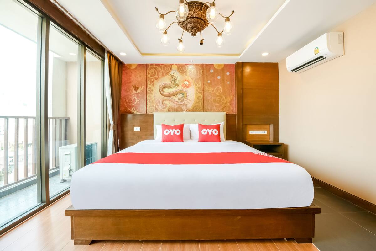 A hotel room with a large bed, a chandelier, a wall-mounted air conditioner, and a view through a large window.