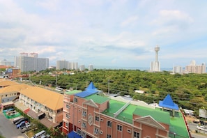A multi-story building with a green roof and blue-topped sections, surrounded by lush greenery and a city skyline in the background. - Grande Caribbean Resort (Pattaya)