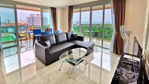 A modern living room with a black sectional sofa, a glass coffee table, and a dining area with blue chairs and a small table. Large windows offer a city view. - Grande Caribbean Resort (Pattaya)