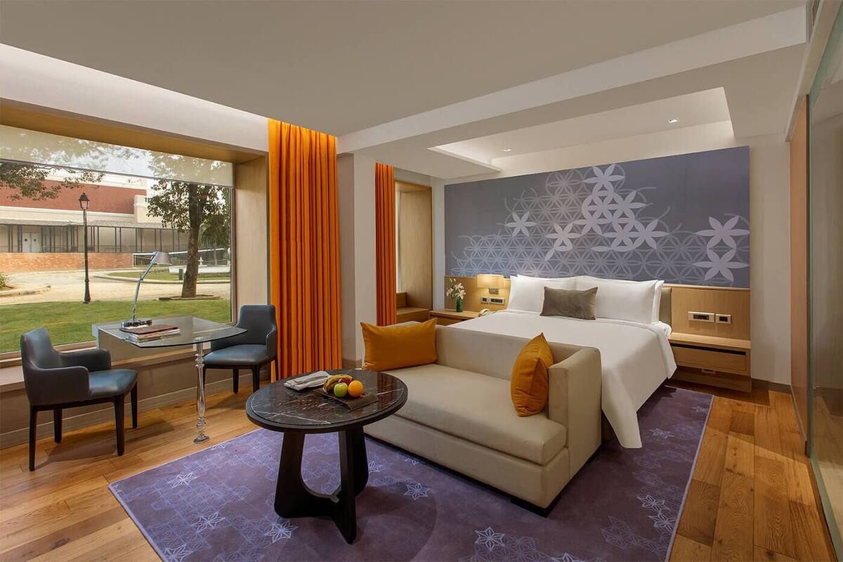 A modern hotel room with a bed, a sofa, a dining table, and a view of the outdoors.