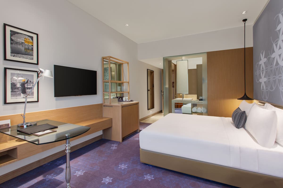 A modern hotel room with a large bed, a desk with a glass top, a flat-screen TV, and framed pictures on the wall.