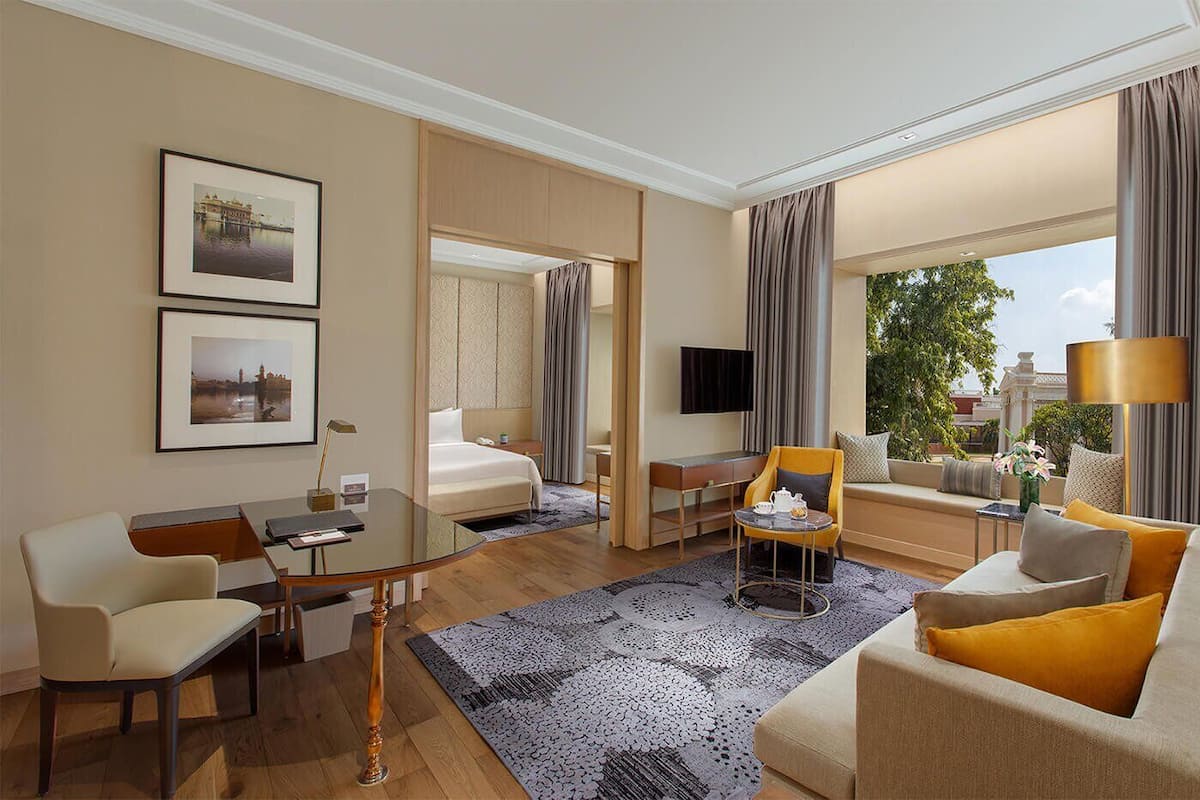 A hotel room with a bed, a desk, a sofa, and a TV. There are two framed pictures on the wall and a window with curtains.