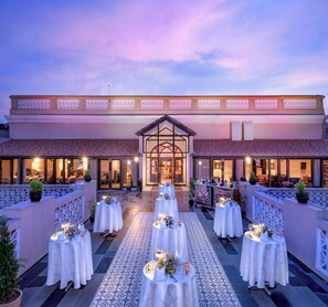 An outdoor banquet hall with multiple round tables set for dining, a central aisle leading to a building entrance, and a decorative pathway. - Welcomhotel by ITC Hotels, Raja Sansi, Amritsar (Ajnala)