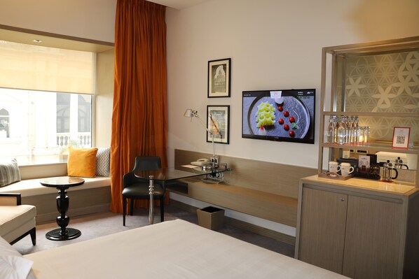 A hotel room with a bed, a small round table, a sofa, a chair, a TV, a minibar, and a window with curtains.