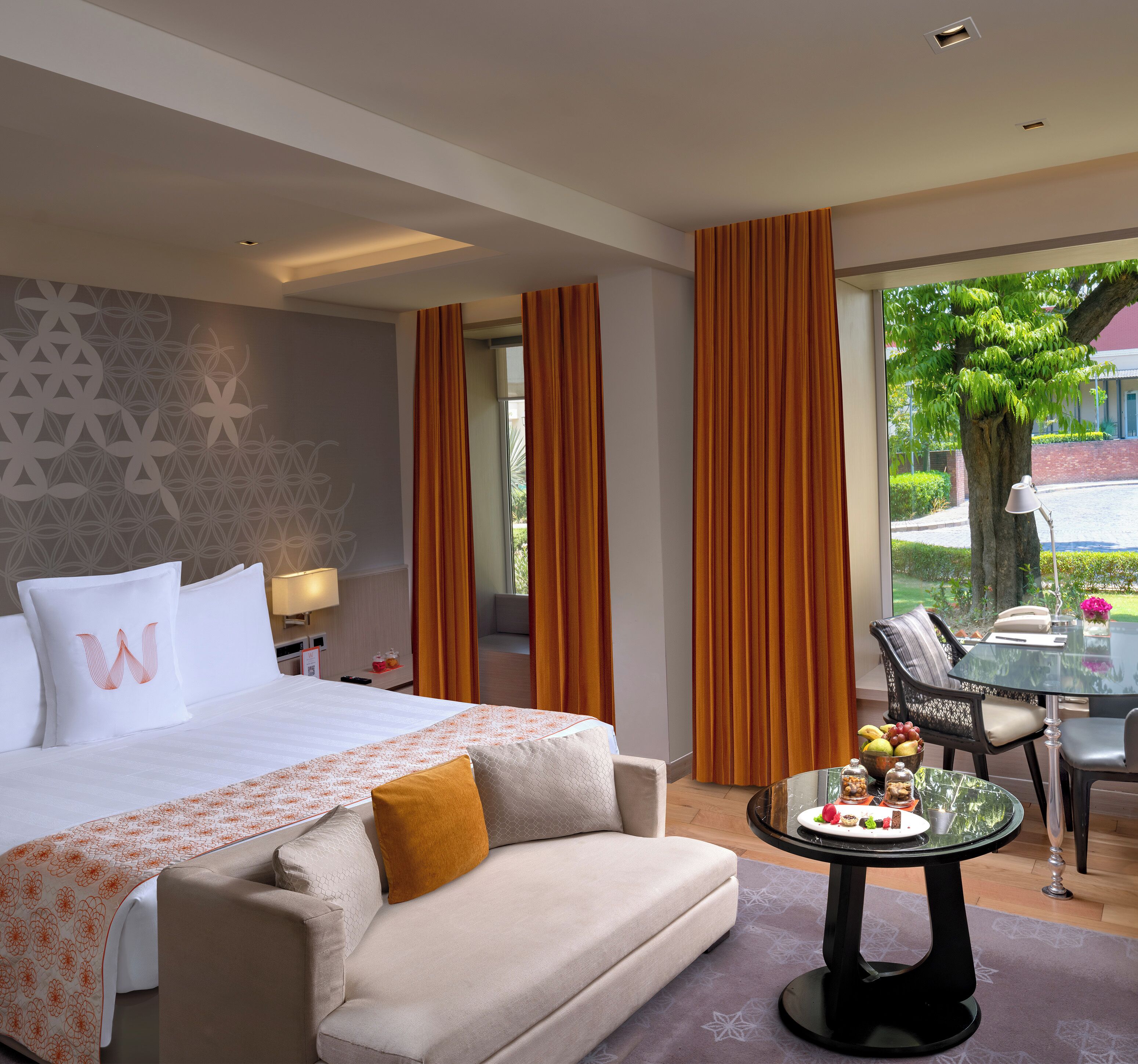 A hotel room with a bed, a sofa, a dining table with a glass top, and a view of the outdoors.
