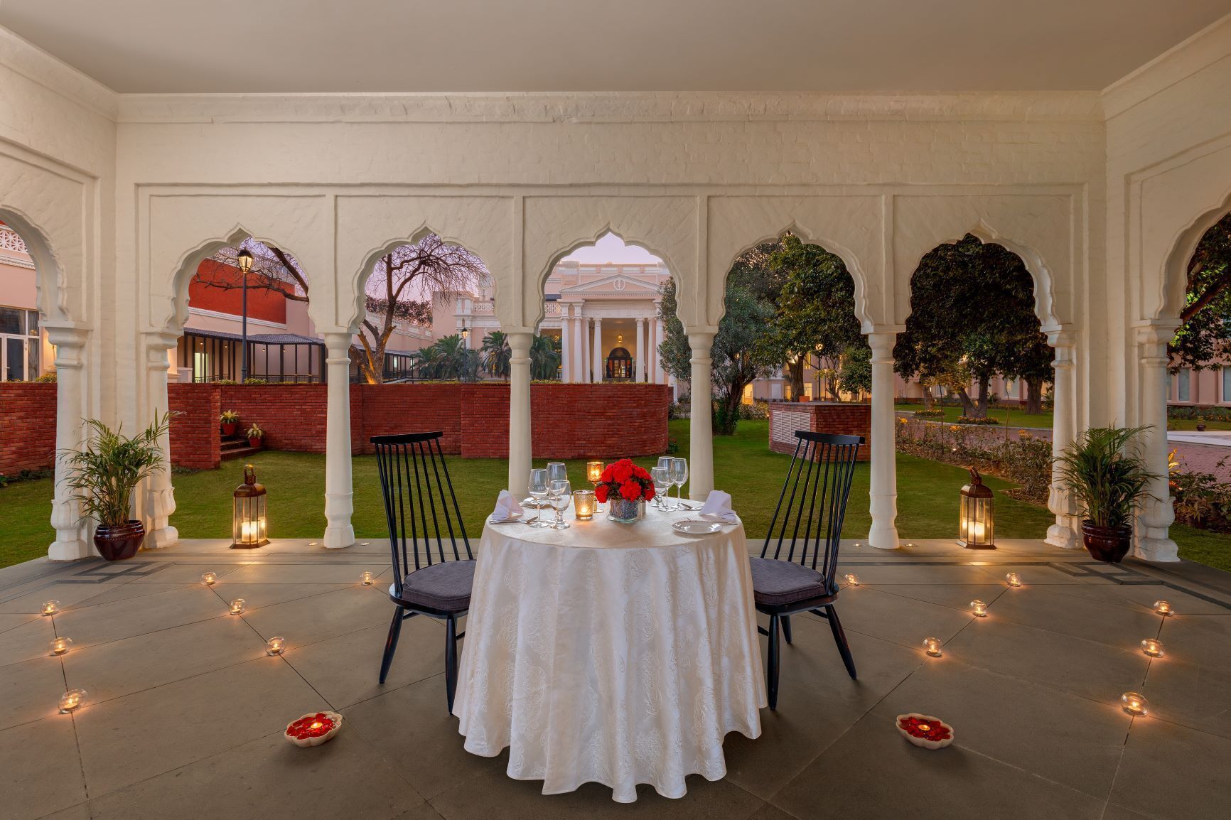 An outdoor dining area with a round table set for two, surrounded by white columns and arches, with a view of a building and greenery in the background.
