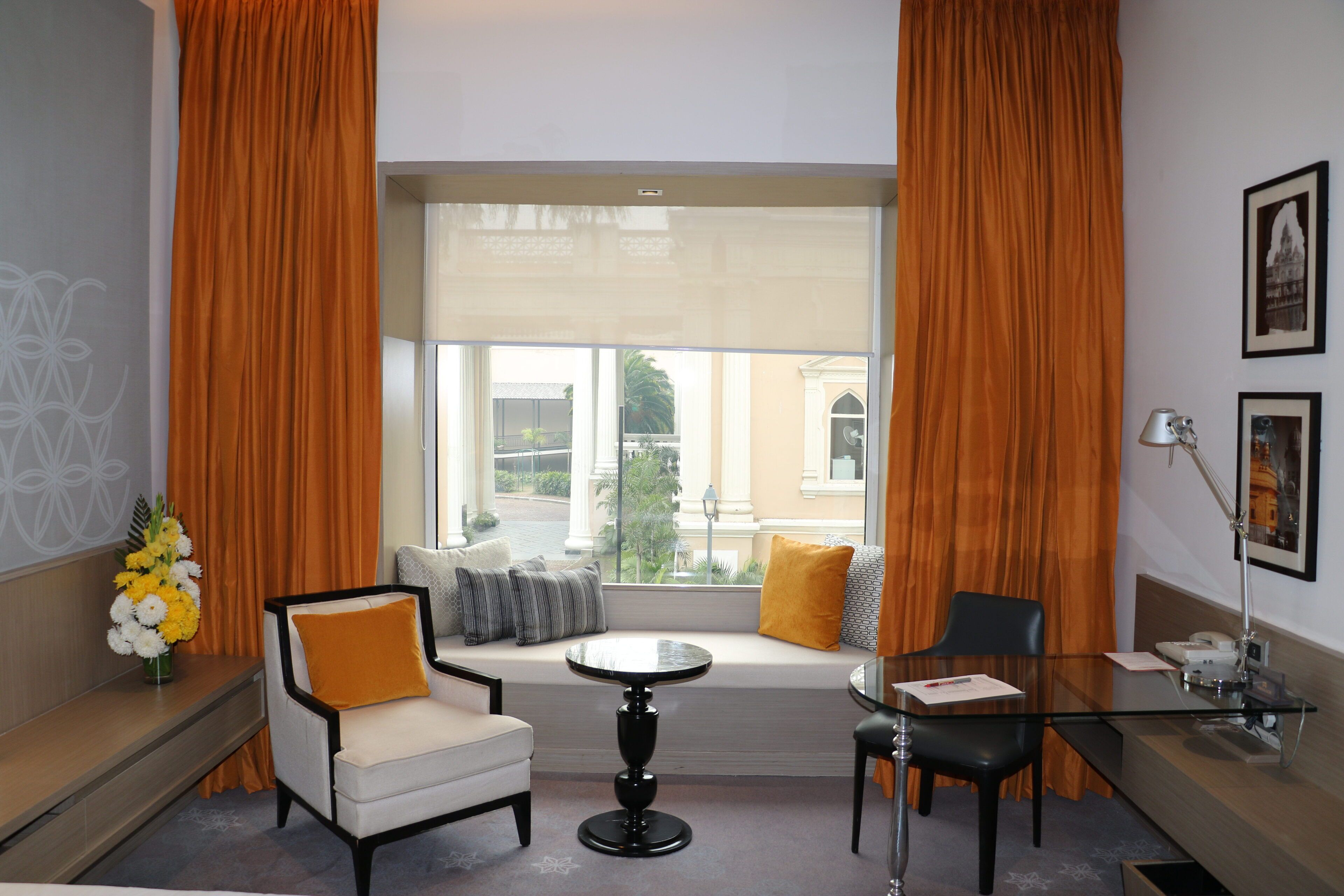 A hotel room with a seating area by a window, a small round table, a chair, a desk with a lamp, and a vase of flowers.